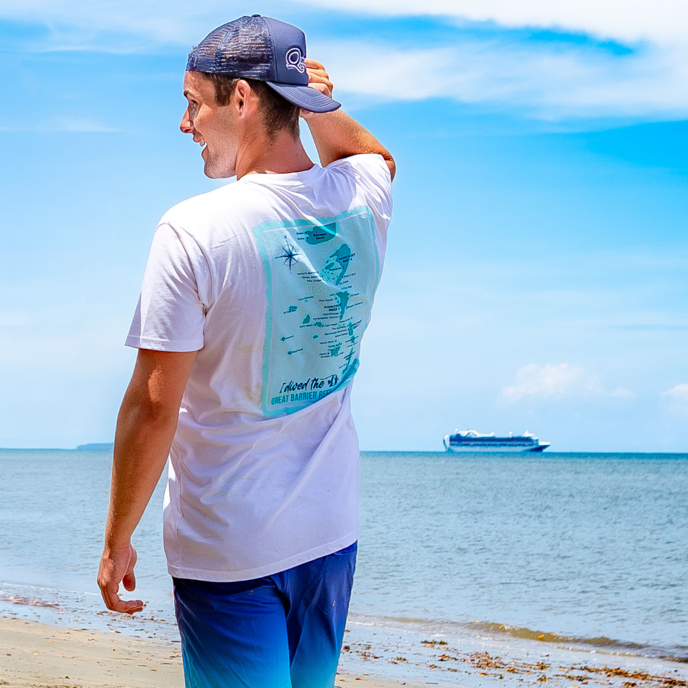 Man on a beach wearing a white t-shirt with a blue graphic and blue shorts, looking at the ocean with a boat in the distance.