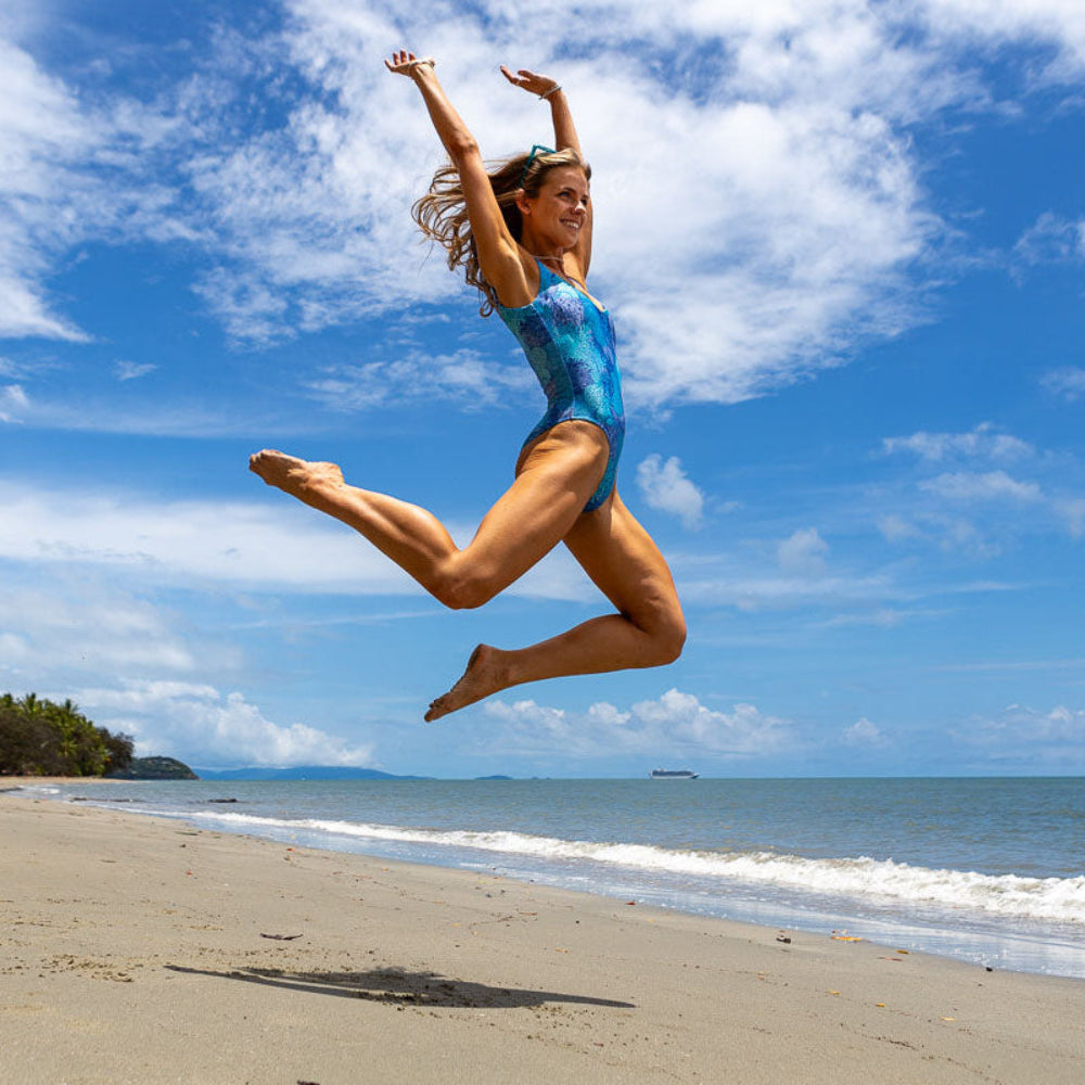 Woman in a blue swimsuit jumping high on a beach with a clear sky.